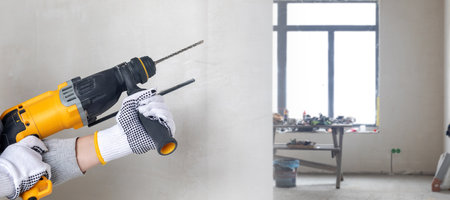In a bright office space, a construction worker in gloves operates a power drill, preparing to make holes in the wall. Tools and equipment are visible in the background, indicating renovation work.の写真素材