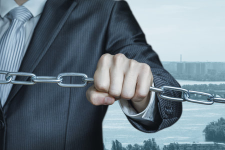 A man in a suit holds a chain tightly, representing the strong connection and unity in business. The background features a cityscape and water, adding depth to the scene.の写真素材
