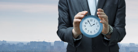 A businessman in a suit stands outdoors holding a clock, emphasizing the importance of time...