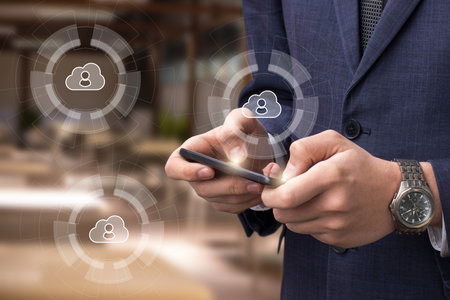 A business professional dressed in a suit interacts with a smartphone, surrounded by digital cloud icons symbolizing connectivity and networking in a sleek office environment.の写真素材
