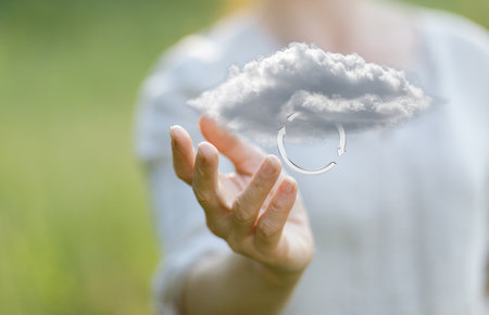 A person extends their hand towards a floating cloud with a circular arrow, symbolizing data processing in cloud computing. The serene outdoor setting enhances the theme of technology and nature.の写真素材