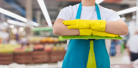 A worker stands confidently in a supermarket, wearing yellow gloves and a blue apron, ready to clean. The bustling environment showcases shelves filled with products and customers shopping.の写真素材