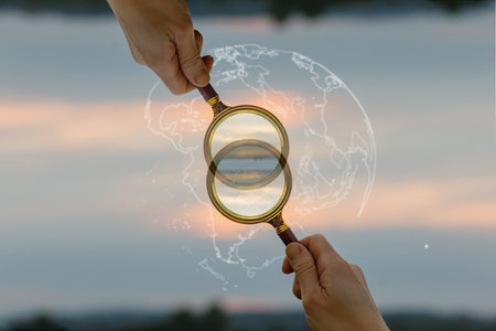 Hands hold a magnifying glass, capturing a stunning sunset view. A faint world map overlays the image, enhancing the theme of exploration and discovery.の写真素材