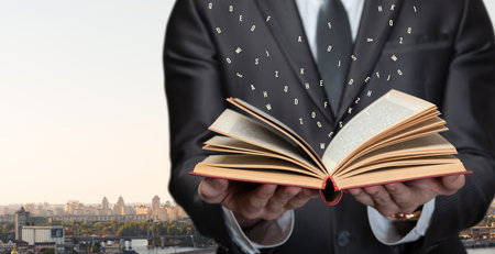 A man in a suit holds an open book with floating letters above it. The city skyline is visible in the background as the sun sets, creating a dramatic atmosphere.の写真素材