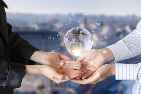 Two hands are positioned together to support a glowing globe, which features symbols of alternative energy, symbolizing global cooperation for sustainability in a modern urban environment.の写真素材