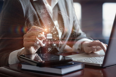 A businessman in a suit interacts with digital icons representing various financial services while working on a laptop. A tablet rests on the desk, indicating a modern workspace.の写真素材