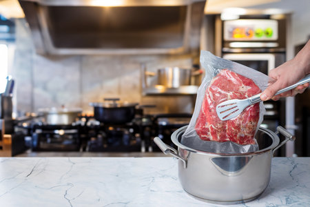 In a modern kitchen, a hand is placing a vacuum-sealed bag of beef into a pot for sous vide cooking. The kitchen features stainless steel appliances and marble countertops.の写真素材
