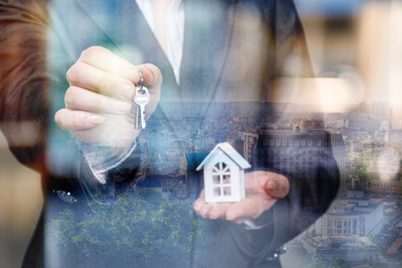A professional man in a suit is holding a key and a small model house. The background shows a vibrant cityscape reflecting his excitement for a new home.の写真素材
