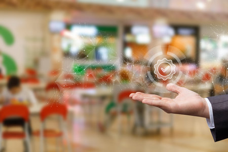 A hand reaches out, showcasing a glowing digital checkmark in a vibrant indoor space filled with people. The atmosphere suggests a focus on quality or approval amidst lively activity.の写真素材