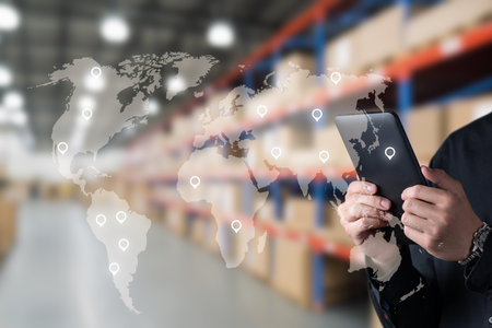 A person is using a smartphone to manage logistics in a warehouse filled with stacked boxes. Digital maps and location pins overlay the scene, illustrating global connections.の写真素材