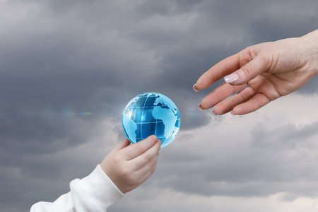 A child's hand reaches out to offer a glowing globe, while an adult hand extends to accept it. Dark clouds in the background show that things are serious and that we need to make a change.の写真素材