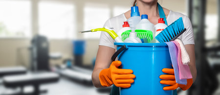 A cleaning professional holds a bucket filled with cleaning supplies and tools. The background shows gym equipment, indicating a focus on maintaining clean fitness spaces.の写真素材