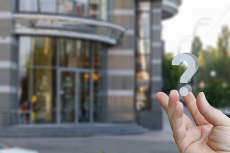 A hand is extended with a transparent question mark positioned above it, in front of a contemporary building. The scene suggests curiosity or inquiry about the location.の写真素材