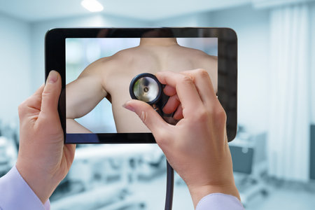 A healthcare professional uses a tablet to examine a patient remotely. The doctor holds the tablet displaying a patient's back while monitoring their health.の写真素材