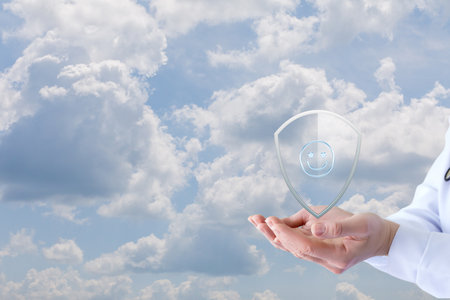 A person holds a transparent shield displaying a smiling face against a backdrop of fluffy clouds, symbolizing health, positivity, and well-being in a serene sky.の写真素材