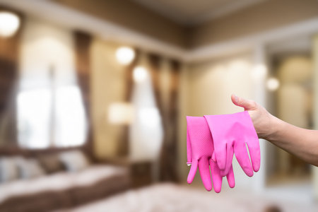 A hand holds bright pink cleaning gloves against a blurred interior setting. The room features large windows and soft furnishings, suggesting preparation for household chores.の写真素材
