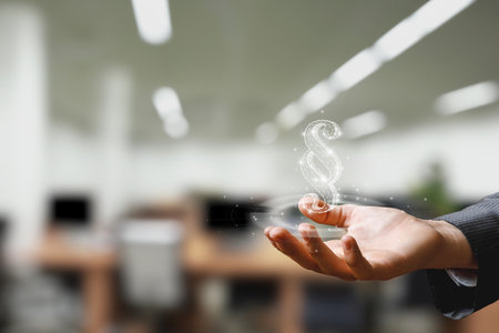 A person in business attire holds a glowing legal symbol in their hand, surrounded by a bright office environment filled with computer desks and blurred coworkers.の写真素材
