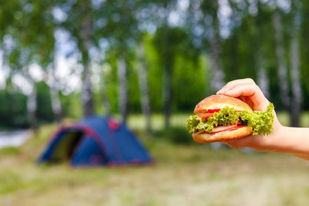 A freshly made burger with lettuce and tomato held in hand while camping near a river under green trees. A colorful tent is set up in the background, enhancing the outdoor vibe.の写真素材