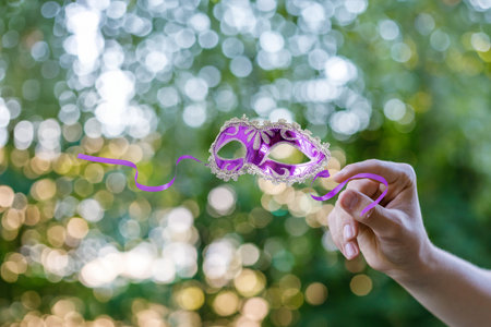 Colorful mask held against a dreamy background outdoors. A vibrant purple mask with intricate details is being held up surrounded by a bokeh of soft greens and lights in a natural setting.の写真素材
