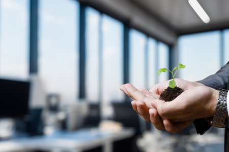 Nurturing new life in a modern office environment. A person holds a small plant in soil symbolizing growth and sustainability in a corporate space.の写真素材