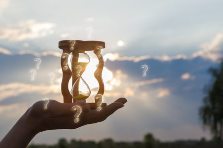 Time and questions beneath a twilight sky. Hand holds an hourglass with flowing sand surrounded by...