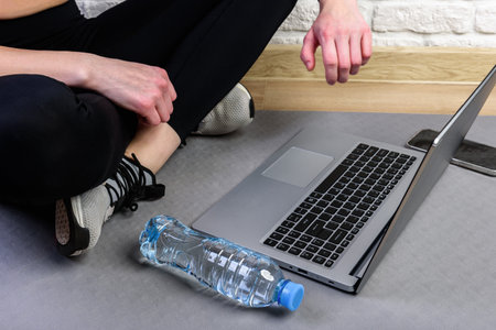 Sporty woman in sportswear is sitting on the floor with dumbbells and bottle of water and is using a laptop at home in the living room.の写真素材