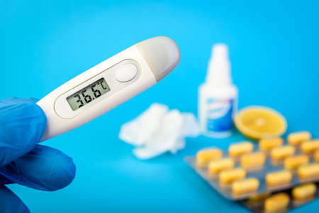 Hands in gloves holding thermometer on blue background with lemon, pills, spray from stuffy nose and pain in a throat. Treatment of colds and flu.の写真素材