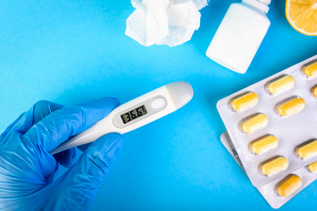 Hands in gloves holding thermometer on blue background with lemon, pills, spray from stuffy nose and pain in a throat. Treatment of colds and flu.の写真素材