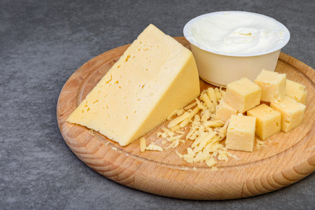 Sliced cheese cubes and cream cheese on wooden board on gray background.