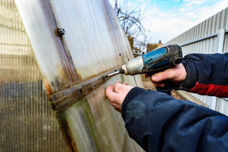 Man inspects old greenhouse for renovation on a spring day, greenhouse repair. Removal old polycarbonate using electric screwdriver.の写真素材