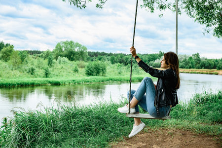 A happy young girl is sitting on a swing in a beautiful tree by the river.の写真素材