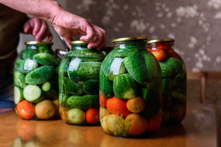 Preservation of seasonal vegetables. A woman pouring the prepared marinade into jars with cucumbers and tomatoes in the kitchenの写真素材