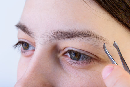 A young girl plucking her eyebrows with tweeters on a gray background