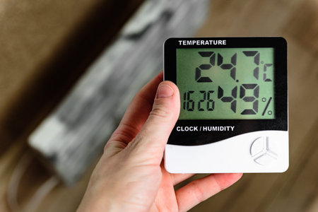 Hand checking room temperature and humidity on a digital thermometer near a heating radiator
