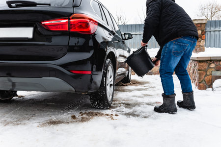 Man hand-washing washing wheels of black car from dirt and salt using a brush and bucket in winter