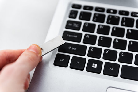 Hand holding silver USB flash drive connected to laptop on table