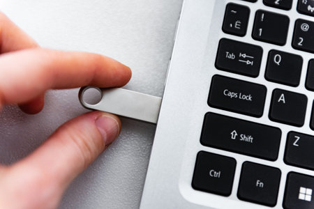 Hand holding silver USB flash drive connected to laptop on table