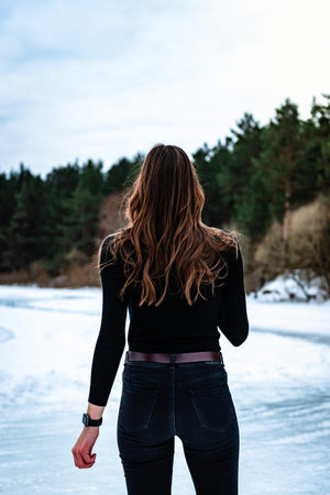Portrait of young girl with long hair in black clothe standing on a frozen lake surrounded by a...