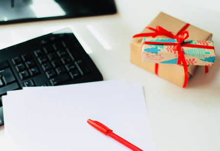 New Year's gifts in wrapping paper with a red ribbon lie on the office table next to the keyboard and a blank sheet of white paper and pen. Christmas. Free space for text.の写真素材