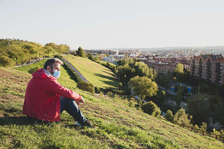 A man in a red windbreaker and a medical mask sits on a green hill and looks at the city skyline....