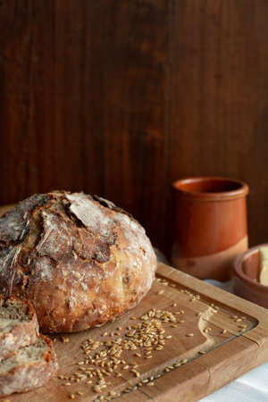 Round shaped homemade bread is on a wooden board where there are cereals and pottery on a wooden...