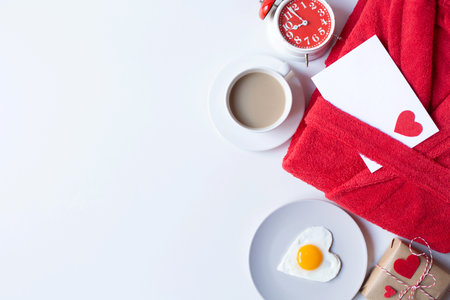 Terry red bath robe, heart-shaped scrambled eggs, a gift, a coffee mug, an alarm clock and a letter envelope are on a white background. Valentine's Day breakfast theme. Free space for text.の写真素材