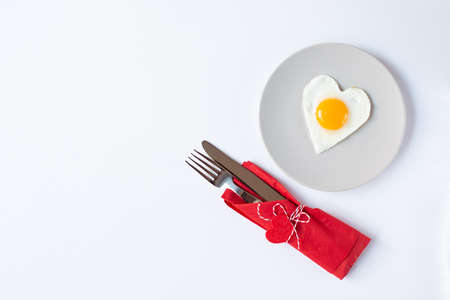 A plate with a fried egg in the form of a heart and cutlery in a red napkin with decorative hearts are on a white background. Valentine's Day breakfast theme. Free space for text.の写真素材