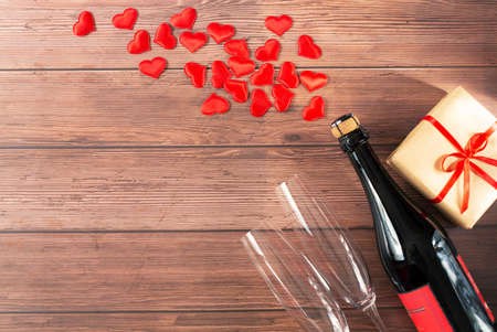 A bottle of champagne, two glass goblets, a gift with a red ribbon, and heart hearts flying out of the bottle are on a wooden background. Valentine's Day theme. Free space for text.の写真素材