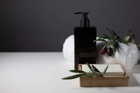 A black jar with a dispenser, a white towel, soap, a sprig of an olive tree with olives are on a gray background. Natural cosmetics theme. Copy space.の写真素材