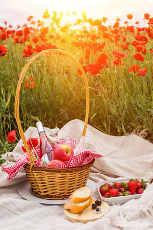 Picnic on a poppy field at sunset. A wicker basket with wine, fruits and berries is on a blanket in a poppy field.の写真素材