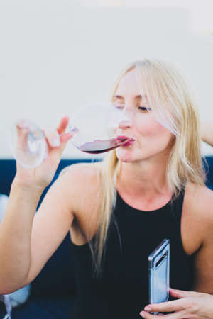 A young girl is drinking red wine from a glass and holding a mobile. The girl at the party drinks...