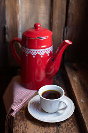 A white cup with black coffee is staning near a red coffee pot on a wooden window sill by the window. Breakfast theme.の写真素材