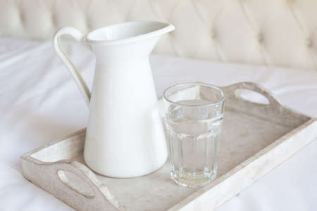A white jug and a glass cup of water stand on a wooden tray on a white bed. Retro style.の写真素材