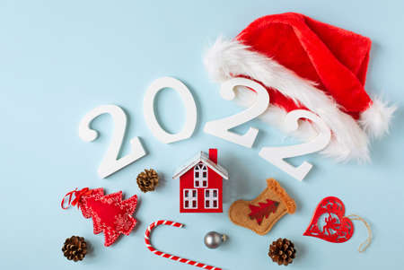 A red decorative house and white numbers 2022 with a red santa claus hat are on a blue background,...